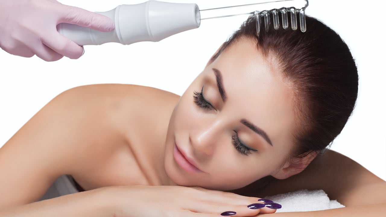 El médico-cosmetólogo realiza el procedimiento Terapia de microcorriente En el cabello de una bella y joven mujer en un salón de belleza.Cosmetología y cuidado profesional de la piel.