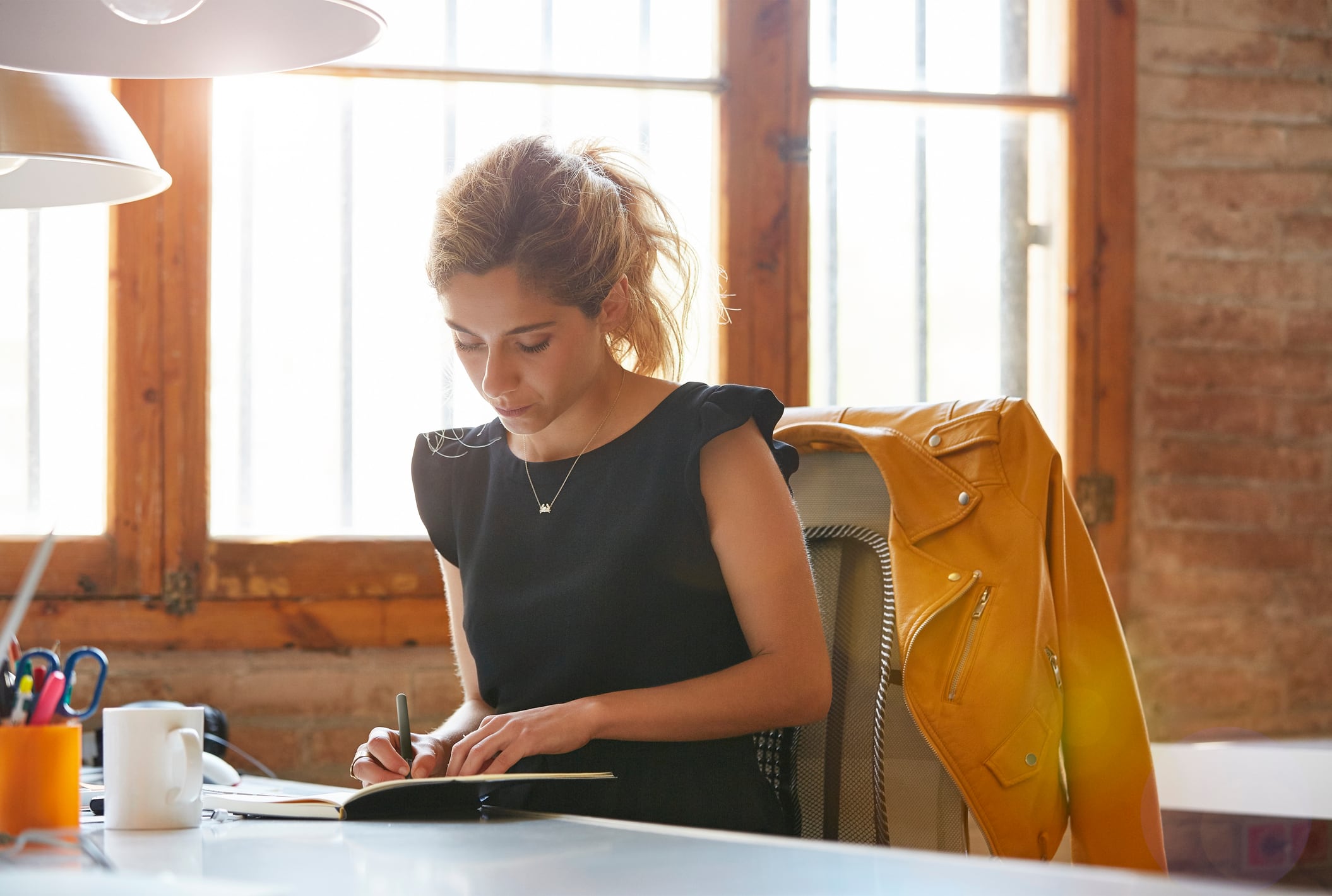 Young businesswoman writing in book at desk. Female professional is sitting on chair. She is making notes in office.