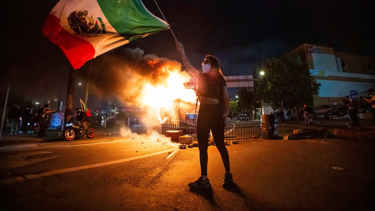 Una mujer ondea la bandera mexicana mientras las llamas brotan de un contenedor de basura en llamas durante una protesta en el centro de Los Ángeles.