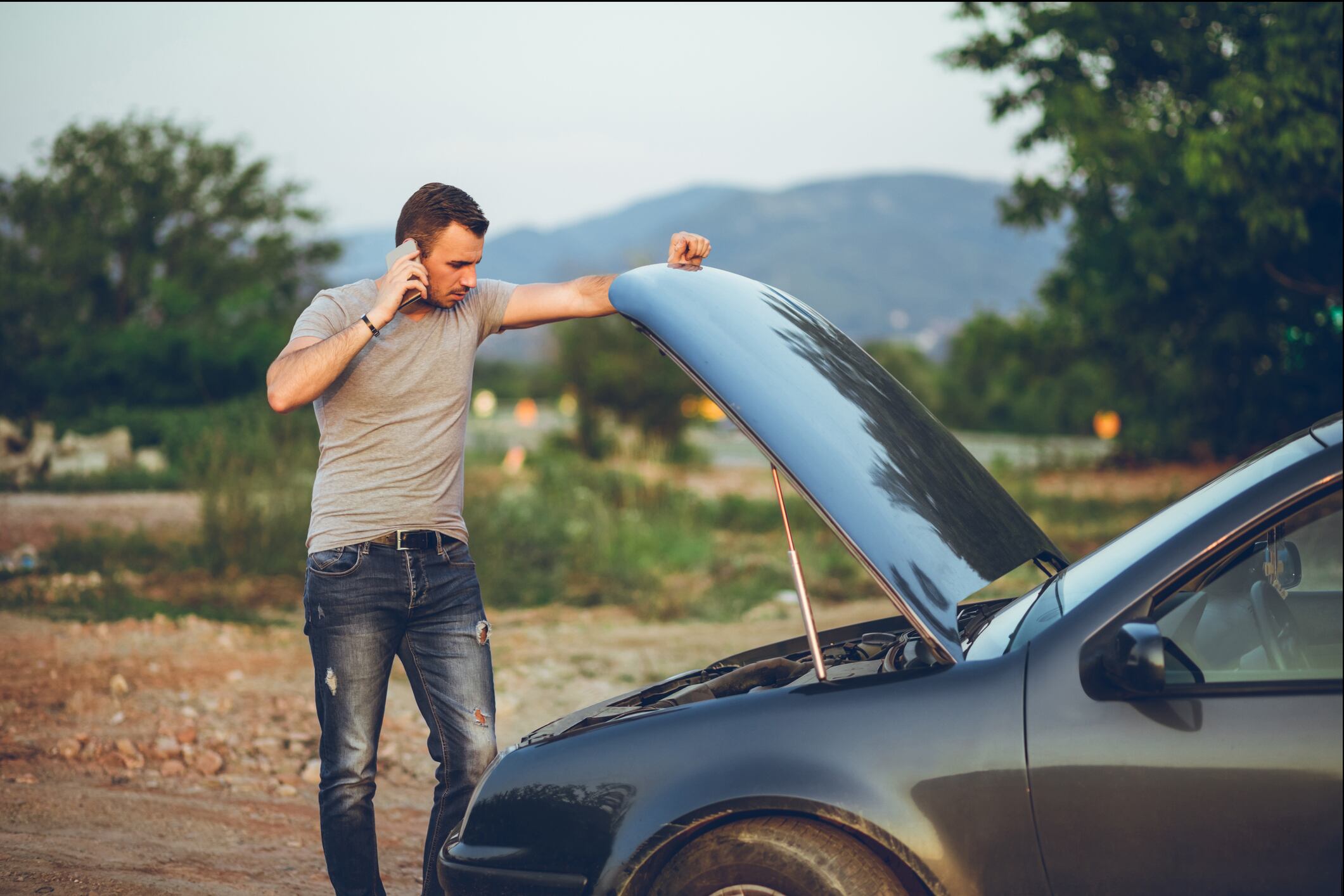 Young man calling for road assistance, standing next to a broken down car