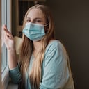 Una mujer joven en pijama con una mascarilla mientras mira por la ventana en casa