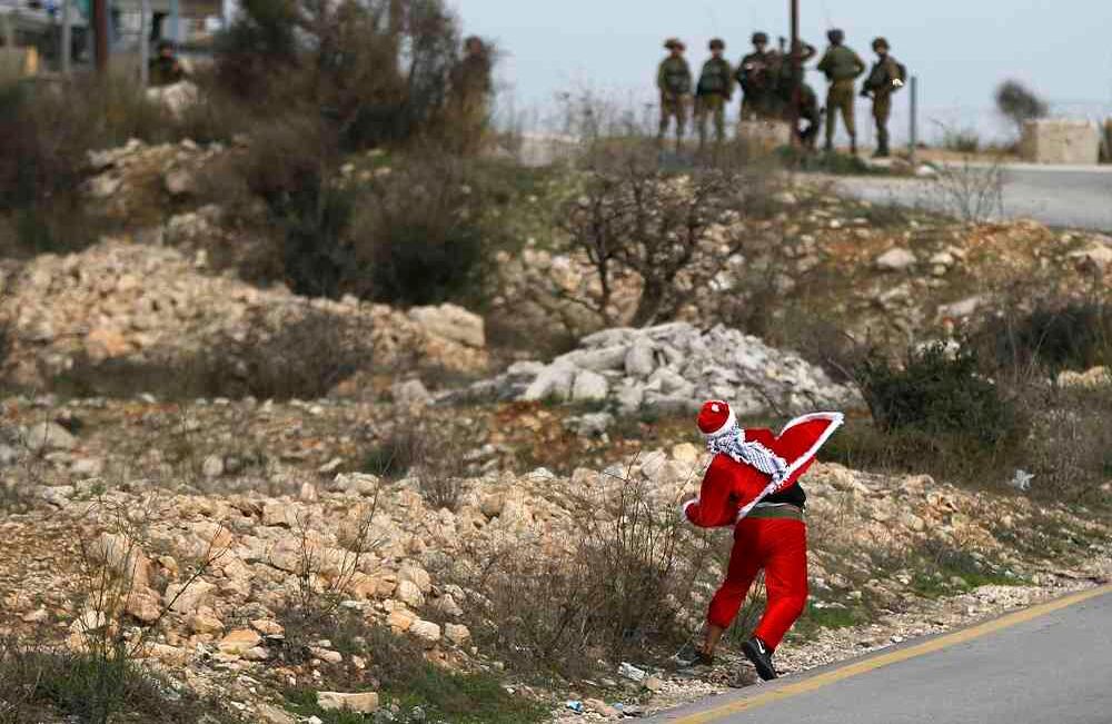 Un manifestante palestino disfrazado de Papá Noel arroja piedras hacia las fuerzas de seguridad israelíes durante los enfrentamientos en el puesto de control de Atarot en las afueras del norte de Jerusalén el 19 de diciembre de 2017. Foto: AFP / ABBAS MOMANI