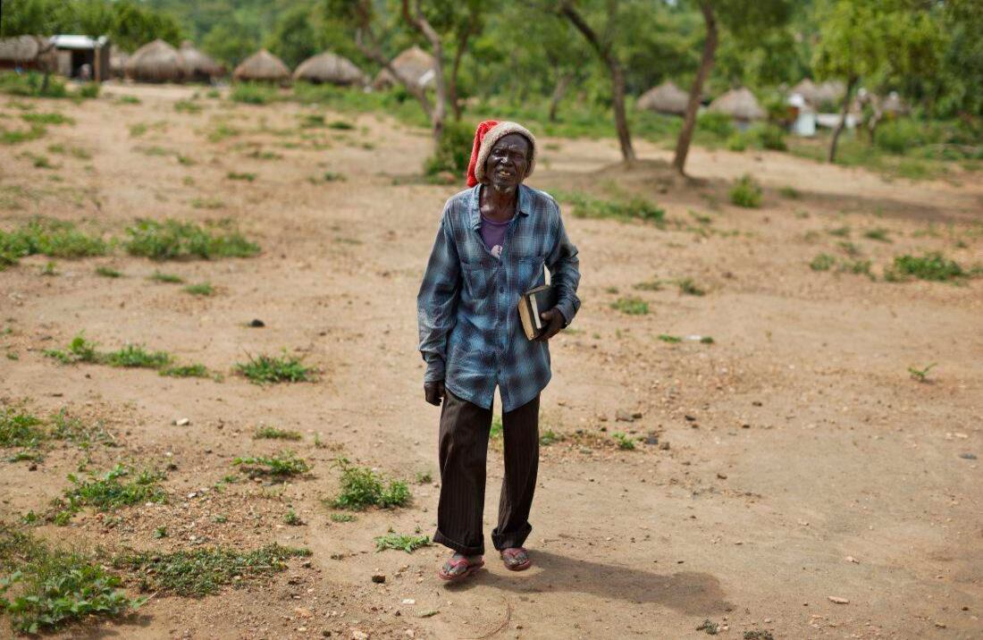 Un anciano sudanés refugiado que lleva un sombrero de Papá Noel y biblias bajo su brazo mientras camina a la iglesia en Bidi Bidi (Foto de AP / Ben Curtis)  