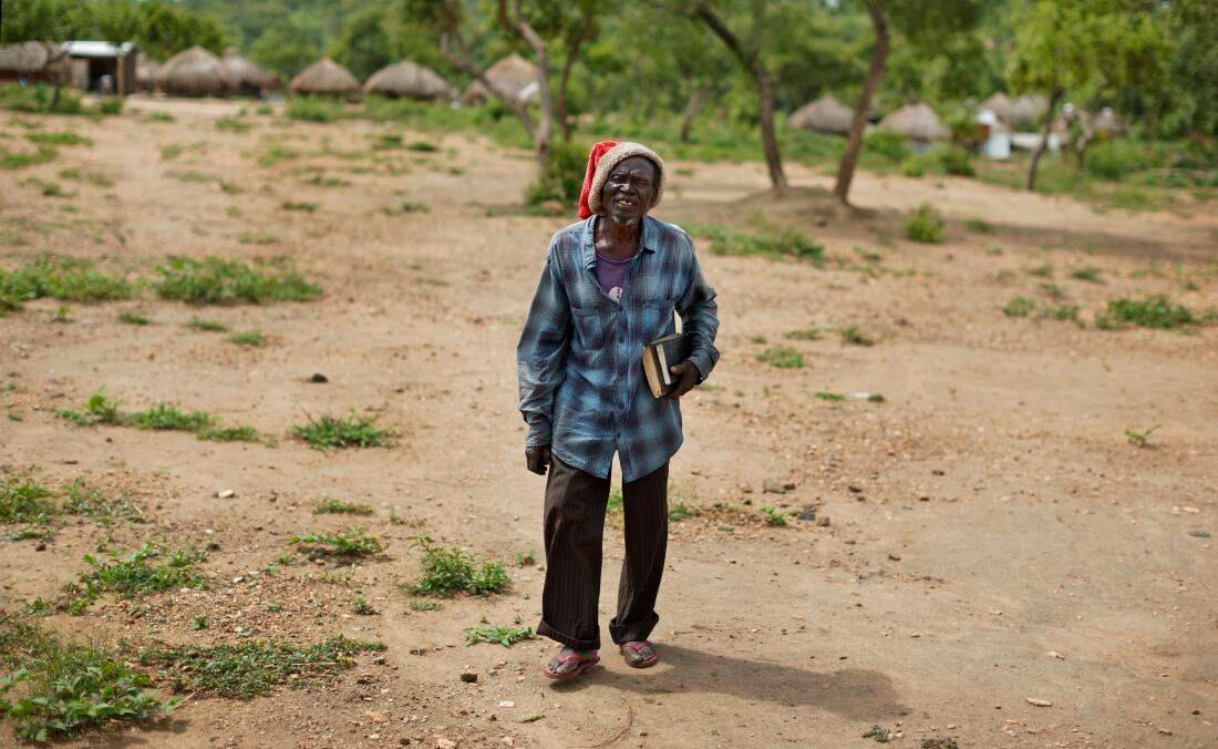Un anciano sudanés refugiado que lleva un sombrero de Papá Noel y biblias bajo su brazo mientras camina a la iglesia en Bidi Bidi (Foto de AP / Ben Curtis)  