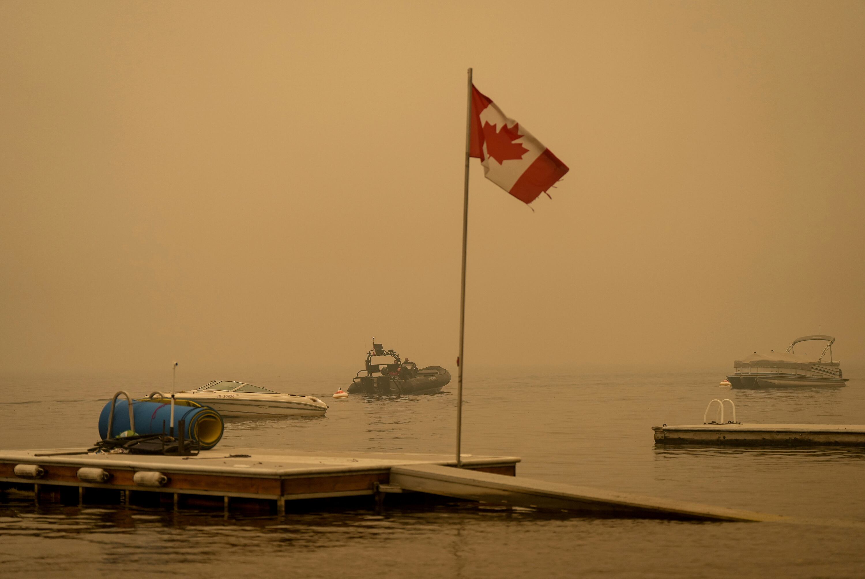 El humo espeso del incendio forestal del lago Lower East Adams llena el aire alrededor de una bandera canadiense ondeando en el viento mientras los oficiales de la Real Policía Montada de Canadá en un barco patrullan el lago Shuswap, en Scotch Creek, Columbia Británica, el domingo 20 de agosto de 2023