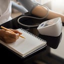 Mujer tomando presión arterial usando esfigmomanómetro digital y grabando en un cuaderno o diario blanco vacío en casa. Concepto médico y sanitario.