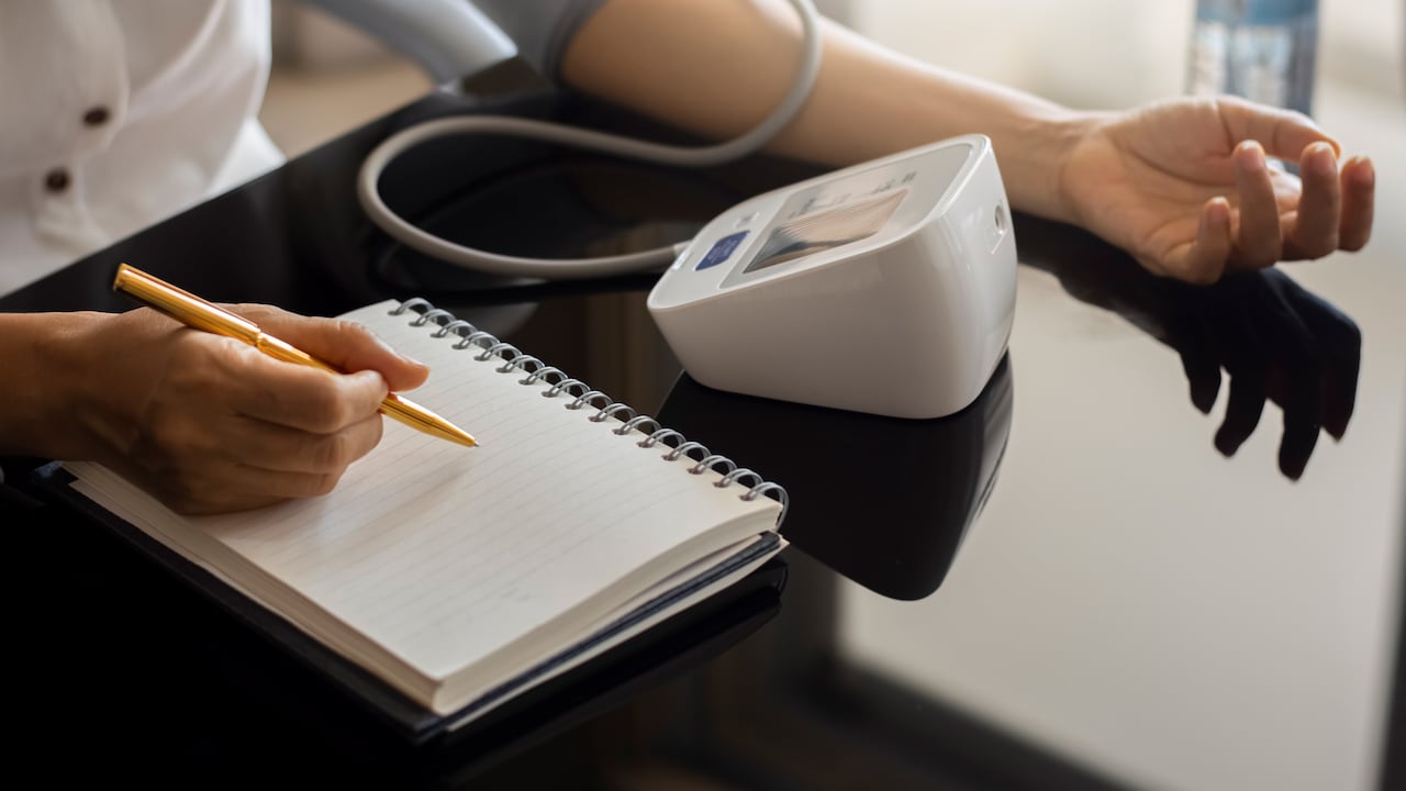 Mujer tomando presión arterial usando esfigmomanómetro digital y grabando en un cuaderno o diario blanco vacío en casa. Concepto médico y sanitario.