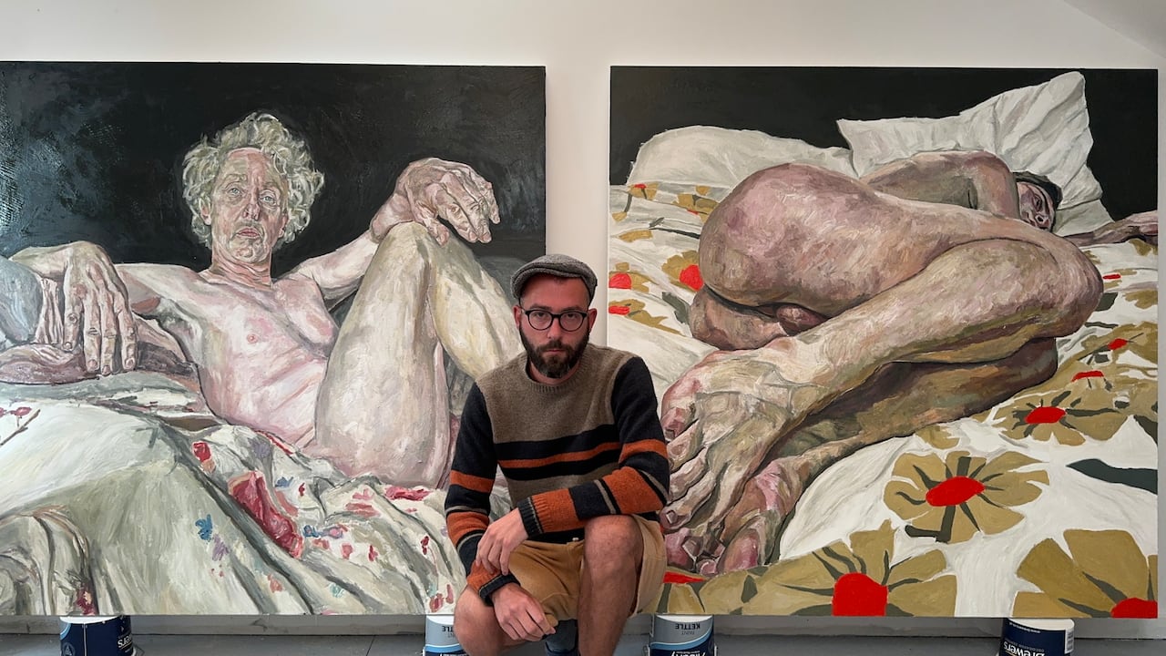 Ian Brennan junto a un dos cuadros con figura humana desnuda, colores terrosos y atmósfera introspectiva.