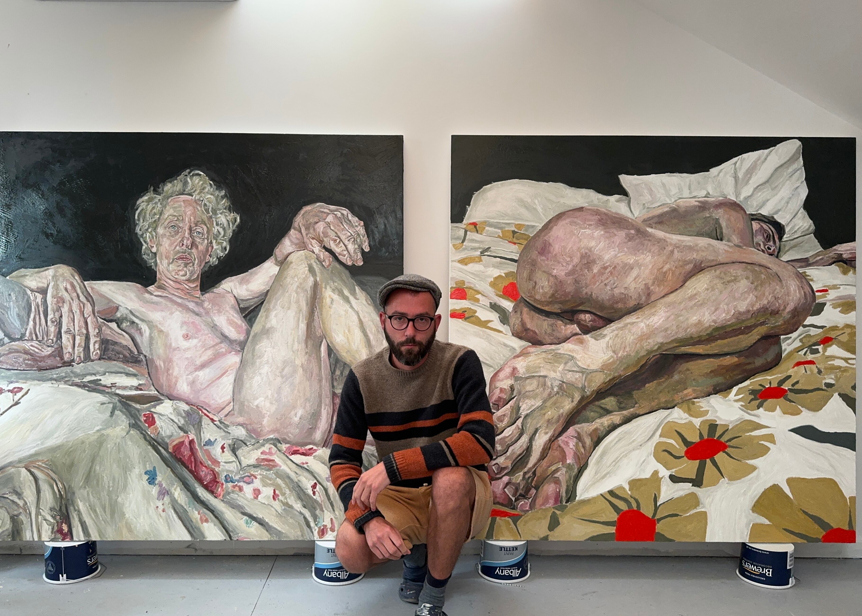 Ian Brennan junto a un dos cuadros con figura humana desnuda, colores terrosos y atmósfera introspectiva.
