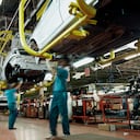 Dos trabajadores vestidos con camisas verdes y pantalones azules trabajan debajo de un automóvil en una fábrica de automóviles. Hay líneas de montaje, automóviles parcialmente construidos, herramientas y equipos eléctricos al fondo.