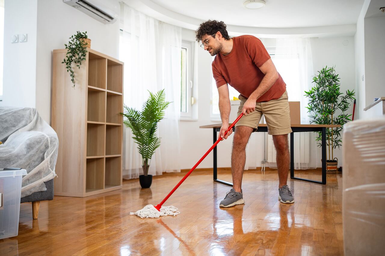 A man in casual attire mopping the shiny wooden floor in a bright, furnished living room, surrounded by modern decor, evoking a sense of cleanliness, organization, and home maintenance.