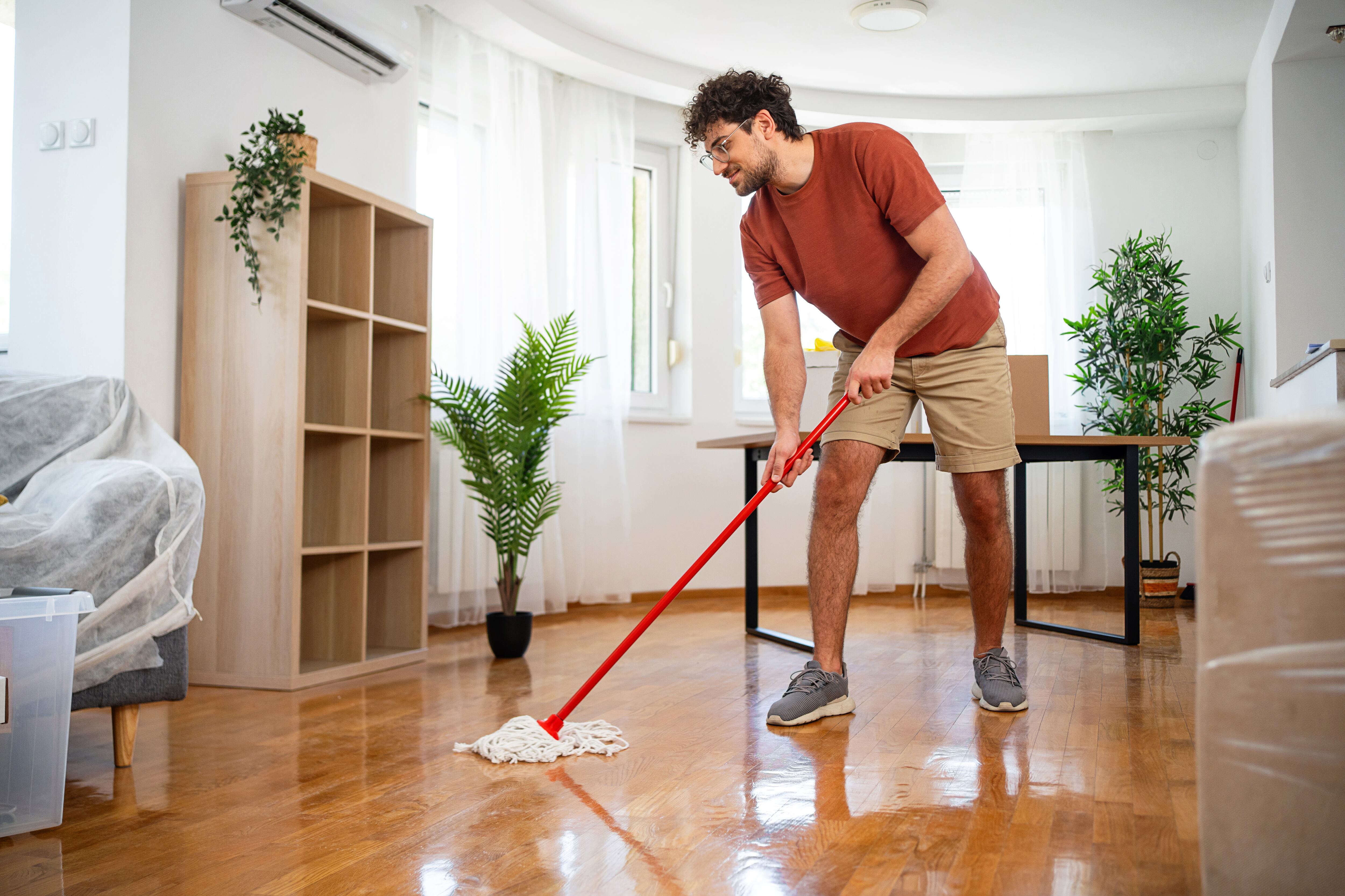 A man in casual attire mopping the shiny wooden floor in a bright, furnished living room, surrounded by modern decor, evoking a sense of cleanliness, organization, and home maintenance.