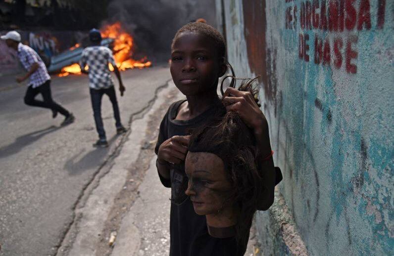 Un niño sostiene una cabeza de maniquí mientras los manifestantes pasan por un auto en llamas durante los enfrentamientos, en el centro de la capital haitiana, Puerto Príncipe, el 12 de febrero de 2019. La imagen corresponde al sexto día de protestas contra el presidente haitiano Jovenel Moise y el uso indebido de los fondos de Petrocaribe. FOTO: HECTOR RETAMAL / AFP