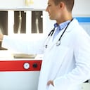 Vista trasera de médicos veterinarios masculinos y femeninos de mediados de los años 30 que examinan rayos X de una extremidad (pata de perro rota). El hombre señala cierta cosa de la pata de este perro mientras una colega sostiene a un paciente peludo. Ambos usan batas de laboratorio blancas.