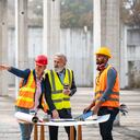 Equipo de proyecto masculino y femenino de pie en la mesa y repasando los planes de construcción y el desarrollador femenino maduro apuntando a algo en el sitio.