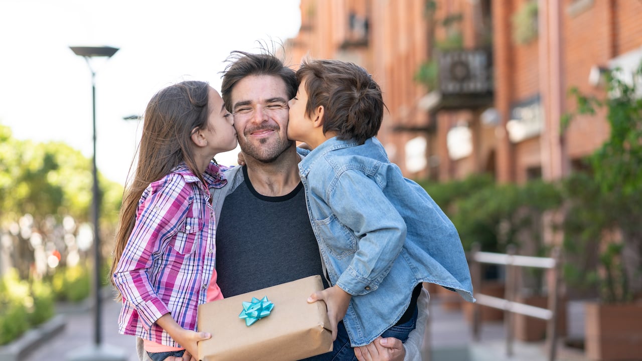 Amoroso hijo e hija besando a papá en la mejilla mientras él los lleva a ambos con un regalo para el día del padre - Conceptos de celebración