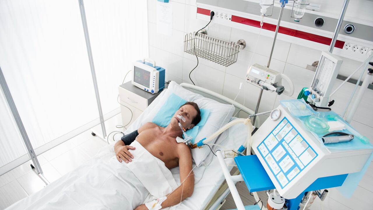 Retrato de vista superior de caballero de mediana edad en ventilador mecánico durmiendo en la sala de recuperación después de la cirugía. El hombre está acostado con goteo intravenoso en su brazo y oxímetro de pulso en el dedo