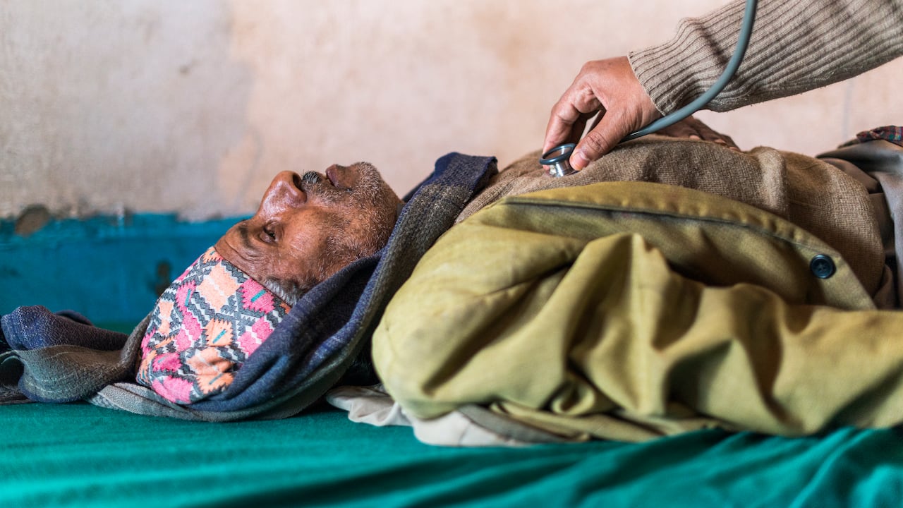 A un anciano que yace boca arriba en un pequeño hospital de Nepal un médico le escucha el latido del corazón con un estetoscopio.