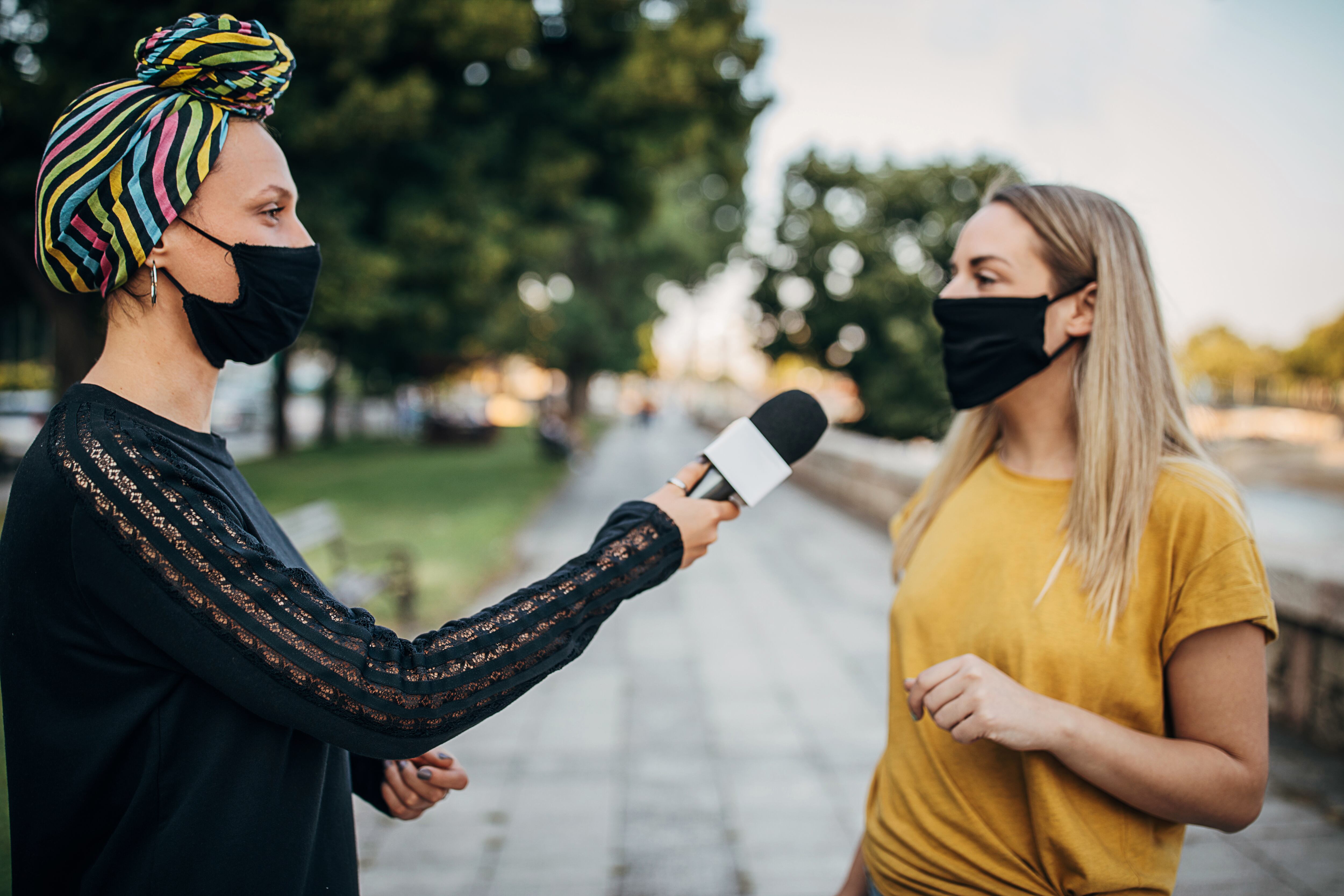 Reportero de noticias femenino entrevistando a una mujer joven en la calle de la ciudad. Ambas mujeres llevan mascarillas protectoras para prevenir la propagación del coronavirus.