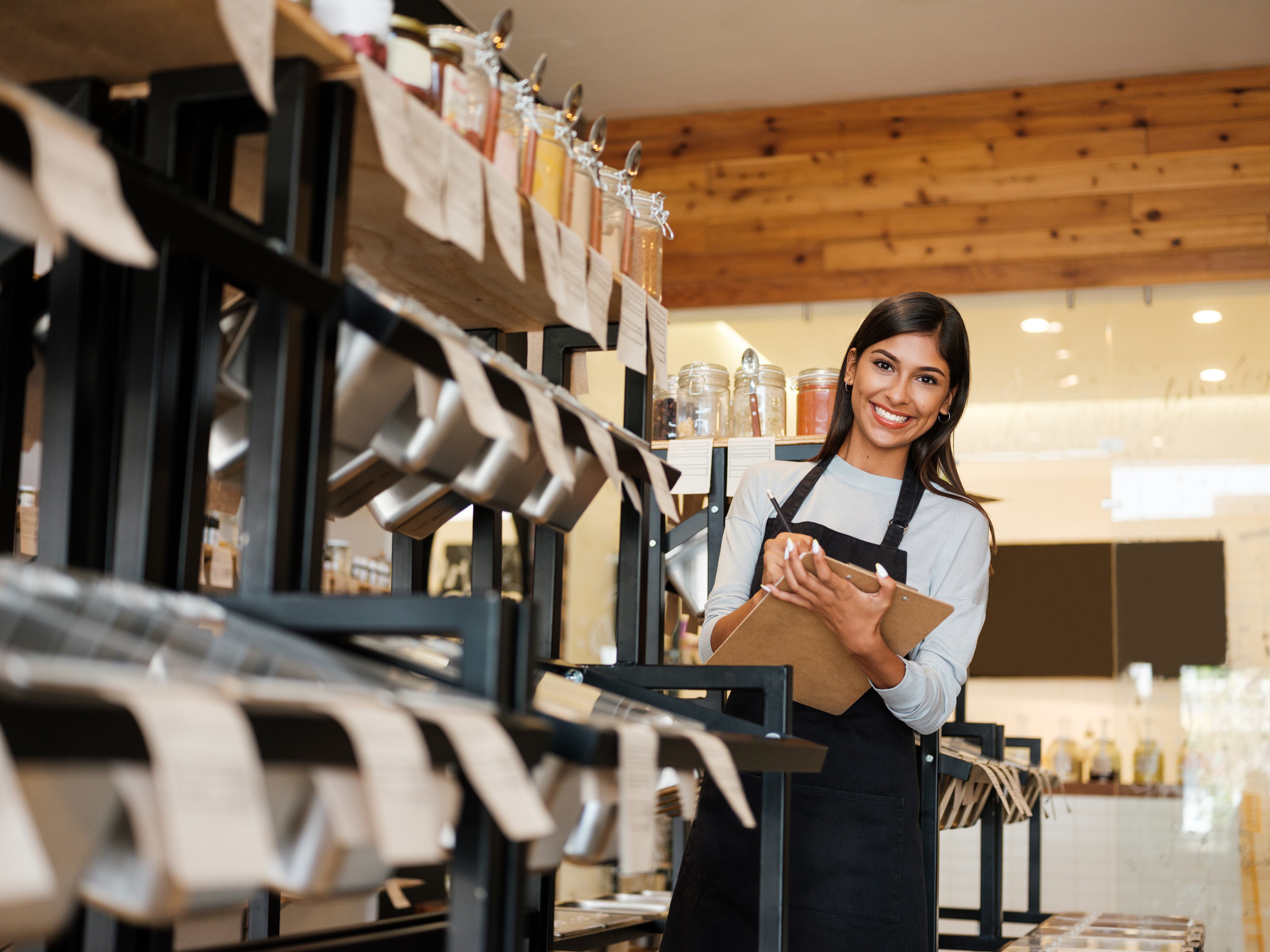 Una empleada latina feliz que trabaja en una tienda ecológica, sosteniendo un portapapeles y sonriendo a la cámara.