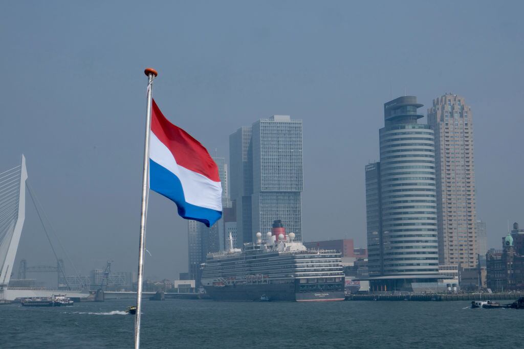 Detrás de una bandera holandesa ondeando, un crucero atraca en el puerto el 2 de mayo de 2024 en Rotterdam, Países Bajos. Al fondo se ven rascacielos modernos. (Foto de Kaveh Kazemi/Getty Images)