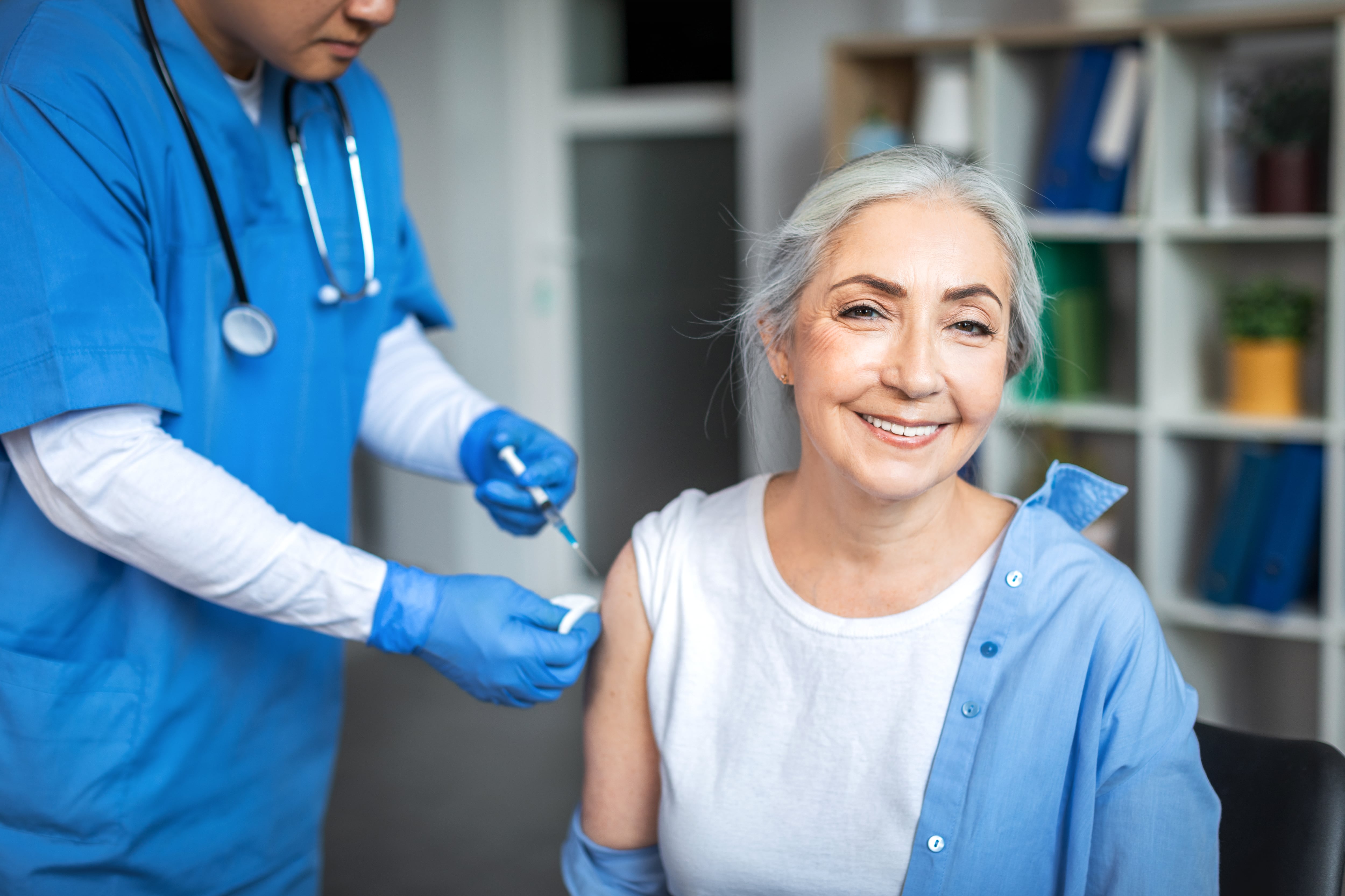 Millennial man doctor make injection to smiling elderly lady patient in clinic office interior. Treatment, medical health care, vaccination and immunization of retired during flu and covid-19 outbreak.
