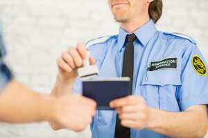 An immigration officer at a country's border is waiting to screen people through the border. He wears a uniform. Here he reads over a person's American passport and gives it an approval stamp and smiles.