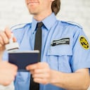 An immigration officer at a country's border is waiting to screen people through the border. He wears a uniform. Here he reads over a person's American passport and gives it an approval stamp and smiles.