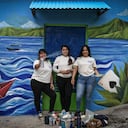Las artistas salvadoreñas Patricia Vega (izq.), Elizabeth Guardado (centro) y Marcela Alvarenga (der.) posan frente a una casa cuya fachada están decorando con el mural «Identidad», en la comunidad 10 de Octubre, en San Marcos, El Salvador, el 2 de octubre de 2026.
Foto: Marvin RECINOS / AFP.