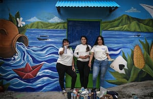 Las artistas salvadoreñas Patricia Vega (izq.), Elizabeth Guardado (centro) y Marcela Alvarenga (der.) posan frente a una casa cuya fachada están decorando con el mural «Identidad», en la comunidad 10 de Octubre, en San Marcos, El Salvador, el 2 de octubre de 2026.
Foto: Marvin RECINOS / AFP.