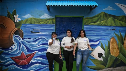 Las artistas salvadoreñas Patricia Vega (izq.), Elizabeth Guardado (centro) y Marcela Alvarenga (der.) posan frente a una casa cuya fachada están decorando con el mural «Identidad», en la comunidad 10 de Octubre, en San Marcos, El Salvador, el 2 de octubre de 2026.
Foto: Marvin RECINOS / AFP.
