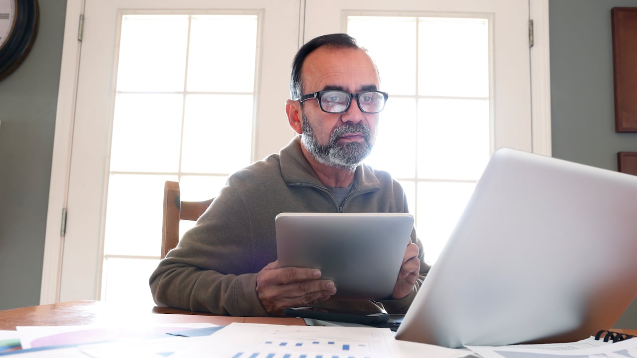 A Hispanic man in his mid fifties sits at his dining room table and peers through his glasses while he holds a digital tablet and looks at his laptop computer. The table is strewn with his work as the morning sun streams in from outside and through the French door window panes in the background and envelopes the room with a bright glow.