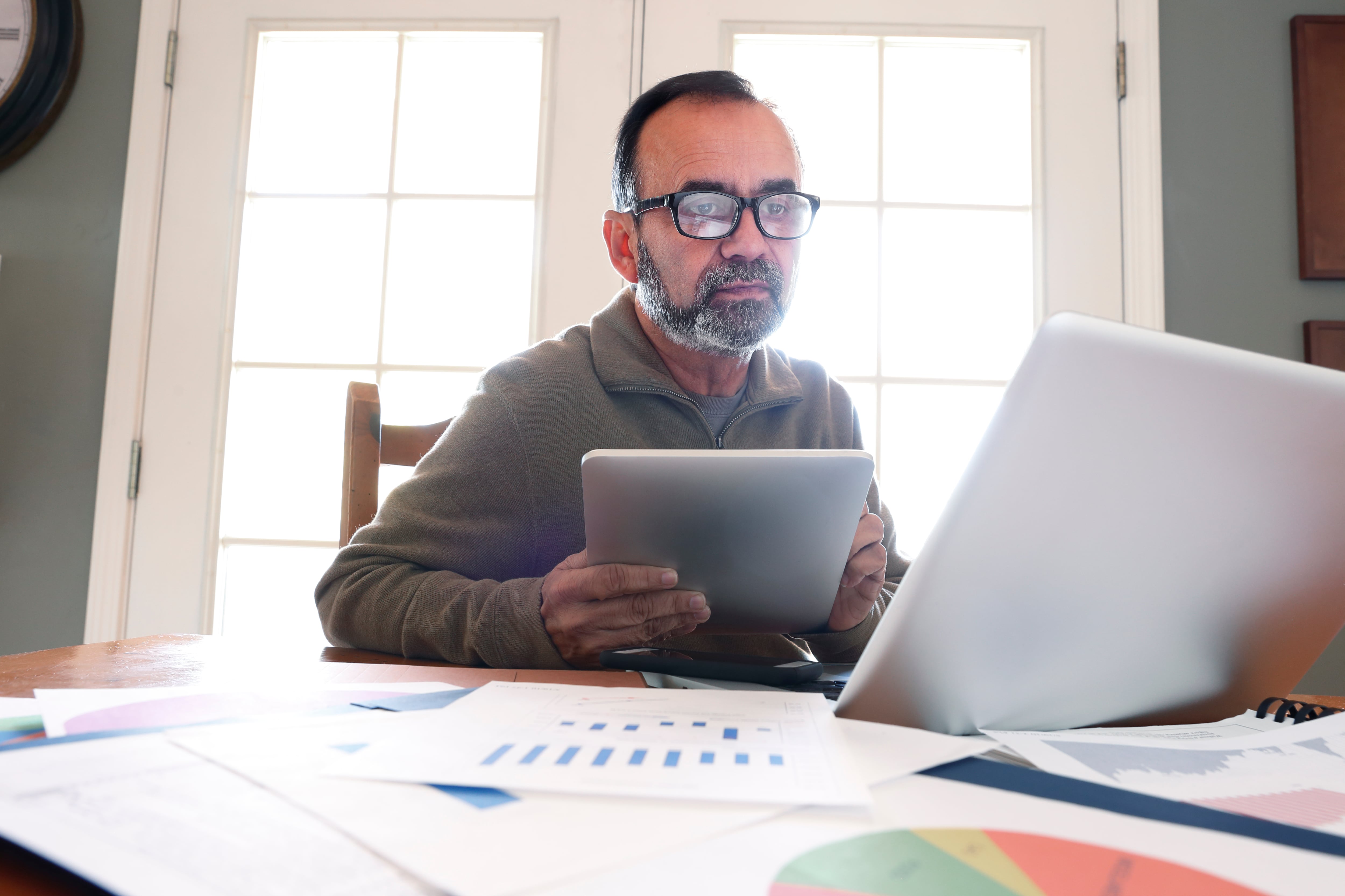 A Hispanic man in his mid fifties sits at his dining room table and peers through his glasses while he holds a digital tablet and looks at his laptop computer. The table is strewn with his work as the morning sun streams in from outside and through the French door window panes in the background and envelopes the room with a bright glow.