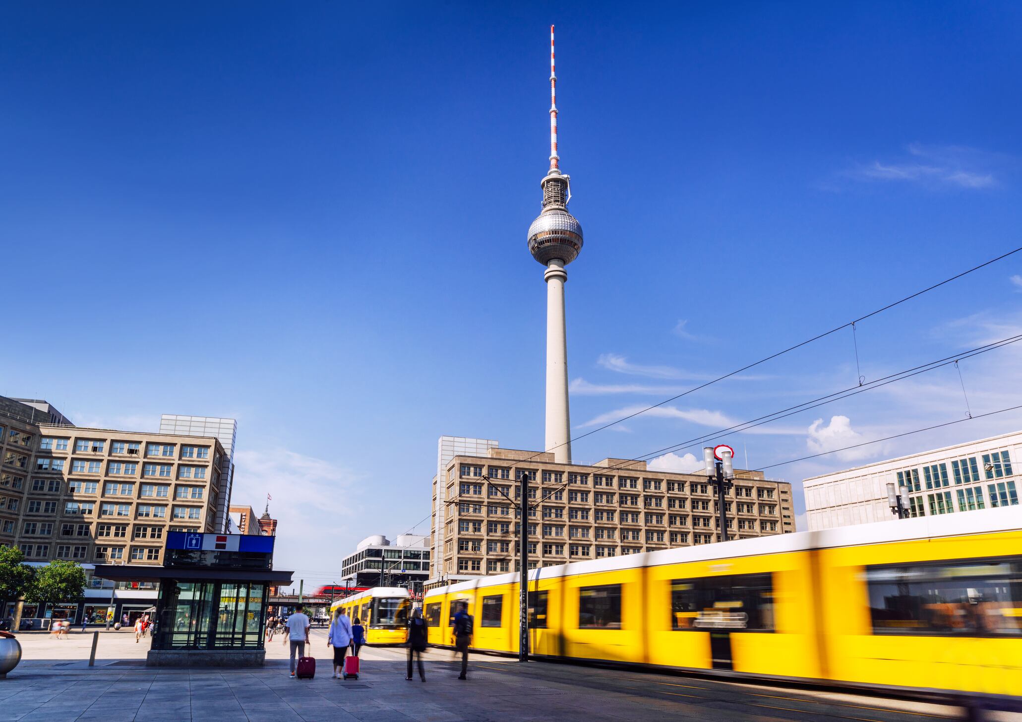 Esta impactante imagen captura una bulliciosa escena berlinesa con la famosa Torre de Televisión, un tranvía amarillo en movimiento y una arquitectura urbana moderna bajo un cielo azul claro.