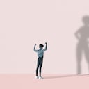 Young woman flexing muscles in front of large superhero shadow on pink background