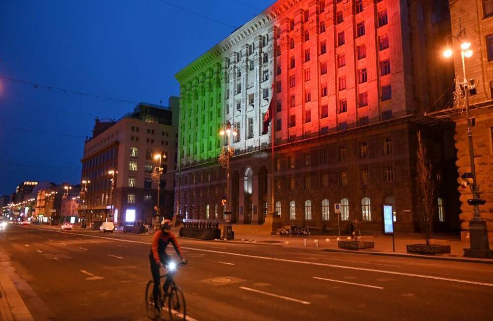 Un ciclista pasa por el ayuntamiento de Kiev, Ucrania, iluminado con los colores de la bandera italiana en solidaridad con el país que actualmente tiene más víctimas fatales por el brote del covid-19. Foto: Sergei Supinsky / AFP