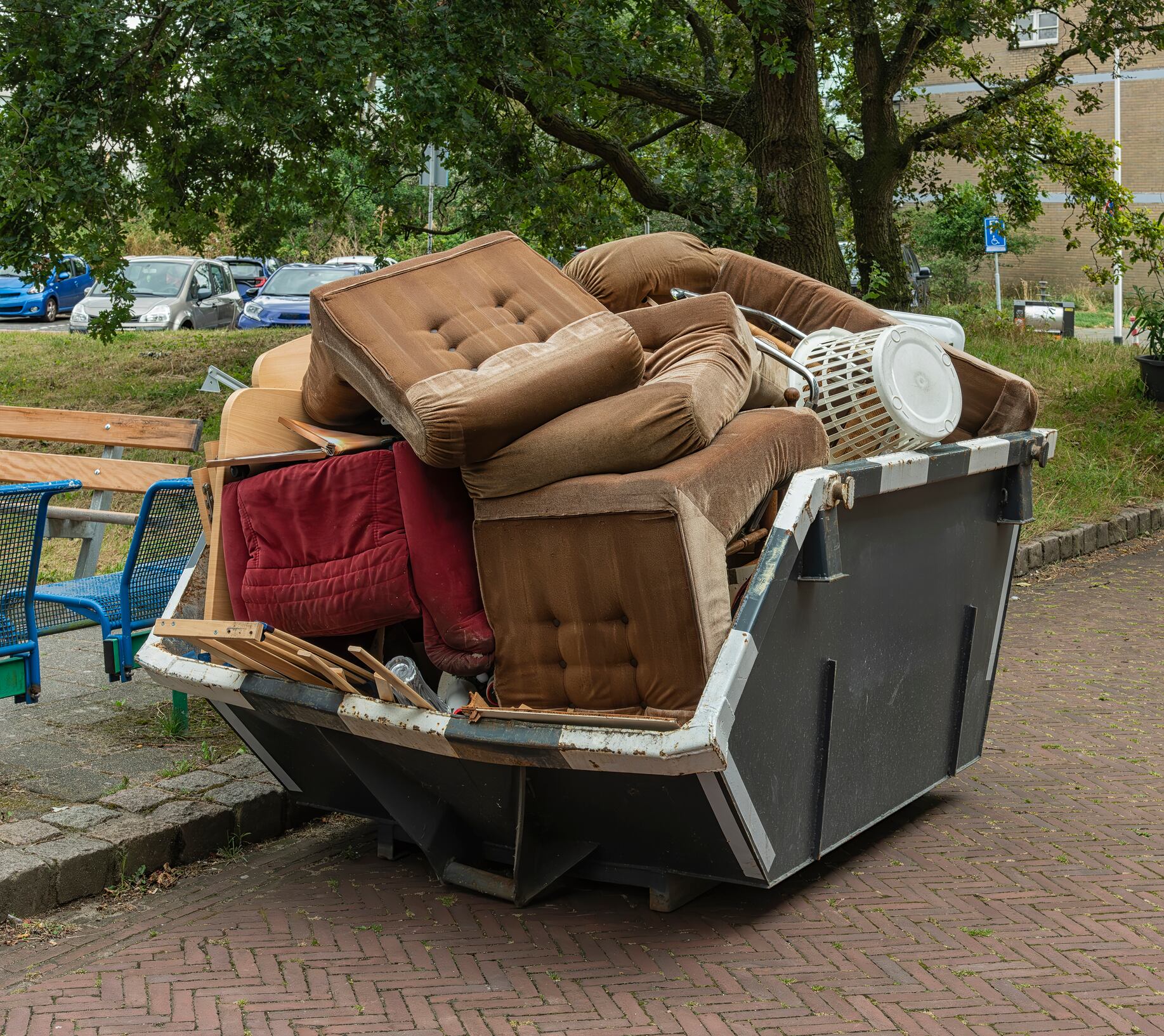 an open steel industrial waste bin filled with chairs and sofas stands outside on the street in a residential area