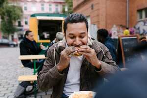 Hombre comiendo una hamburguesa mientras está sentado al aire libre. Foto: Getty Images.