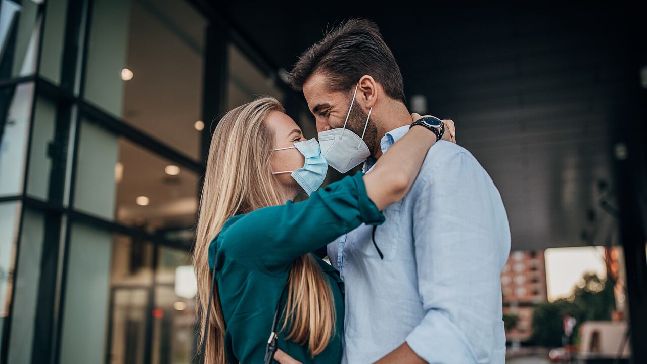 Una joven pareja con tapabocas y besándose al aire libre en la ciudad.