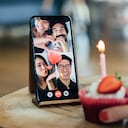 Close-up shot of a birthday cake and a smart phone screen showing international friends having a virtual birthday party on video call.