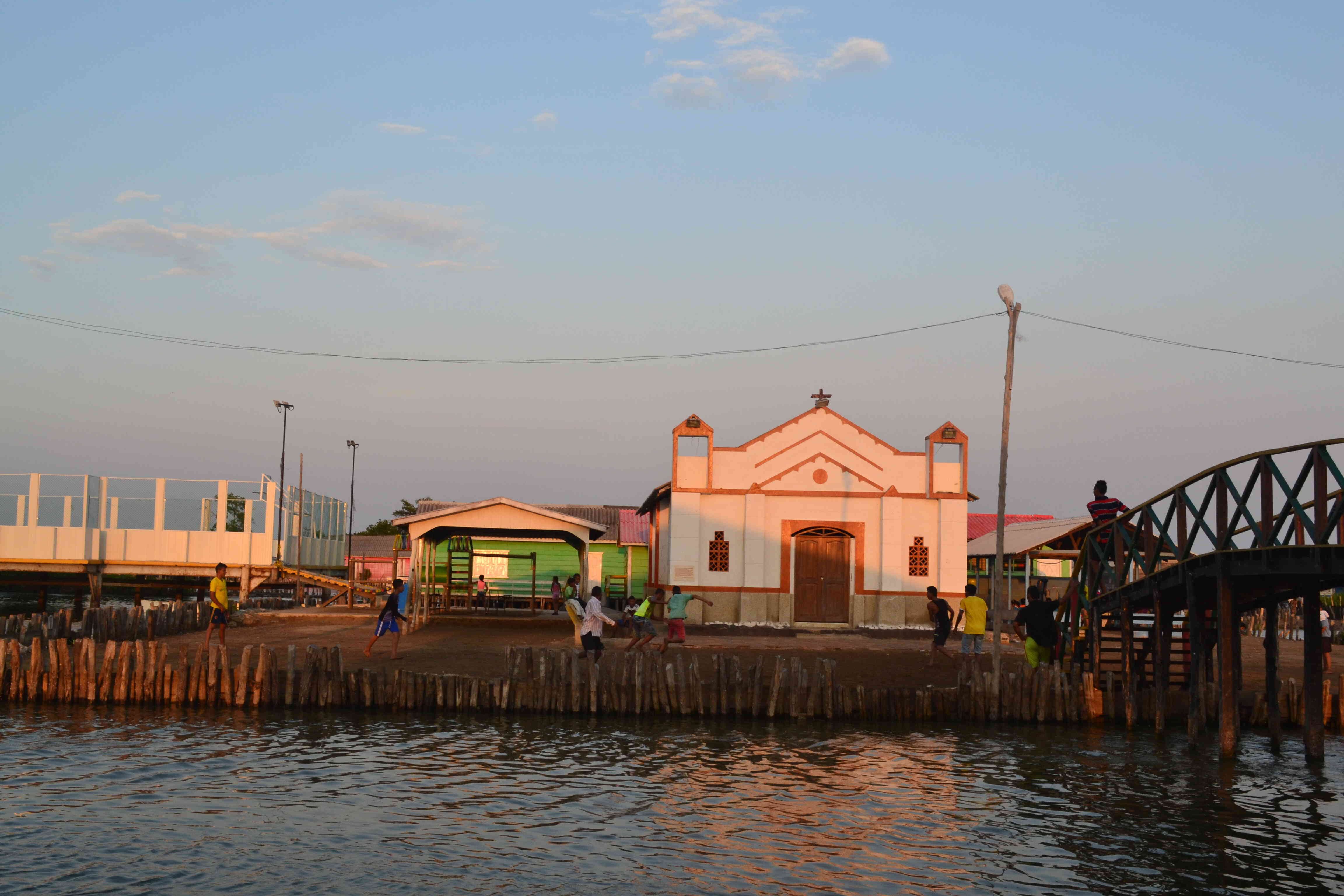 Un grupo de jóvenes juega fútbol en una cancha de tierra frente a la iglesia en Nueva Venecia.