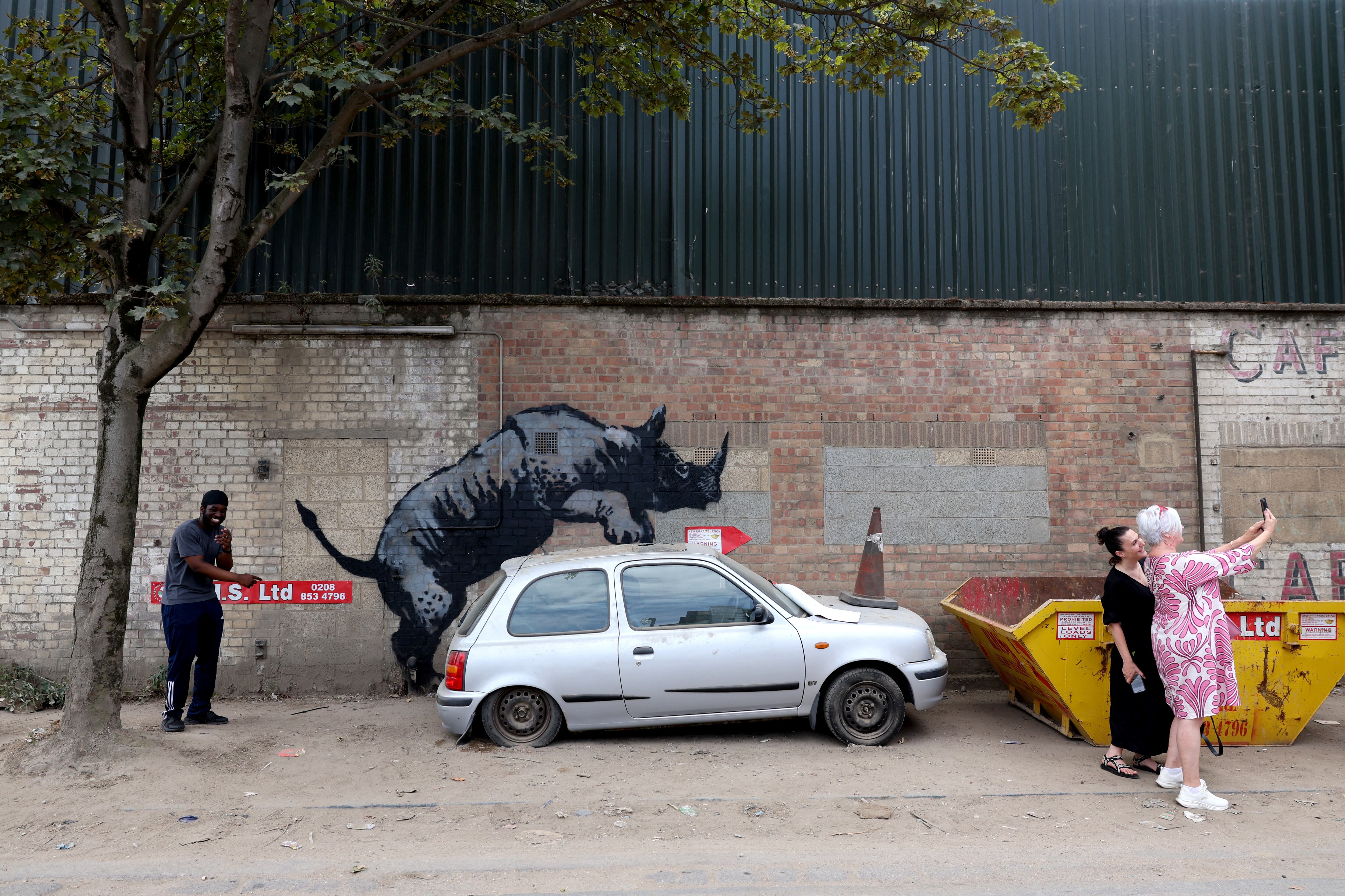 People gather around an artwork by street artist Banksy, the eighth to released in eight days, depicting a rhino mounting a car, on a wall in Charlton, London, on August 12, 2024. The artist confirmed the work to be theirs after posting an image of it on the social media app Instagram. (Photo by Adrian DENNIS / AFP) / RESTRICTED TO EDITORIAL USE - MANDATORY MENTION OF THE ARTIST UPON PUBLICATION - TO ILLUSTRATE THE EVENT AS SPECIFIED IN THE CAPTION