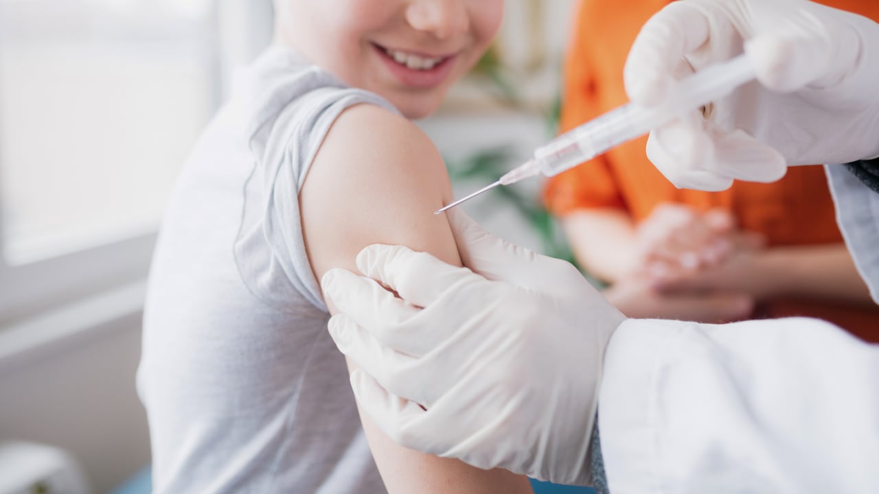 Cerca de un niño caucásico sonriente llevado a una clínica médica por su madre y recibiendo la vacuna Covid 19 en su hombro por un médico varón caucásico