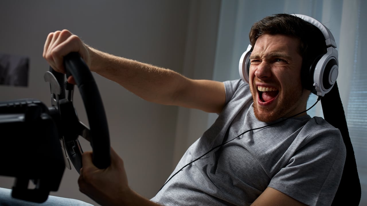 technology, gaming, entertainment and people concept - young man in headphones with pc computer playing car racing video game at home and steering wheel