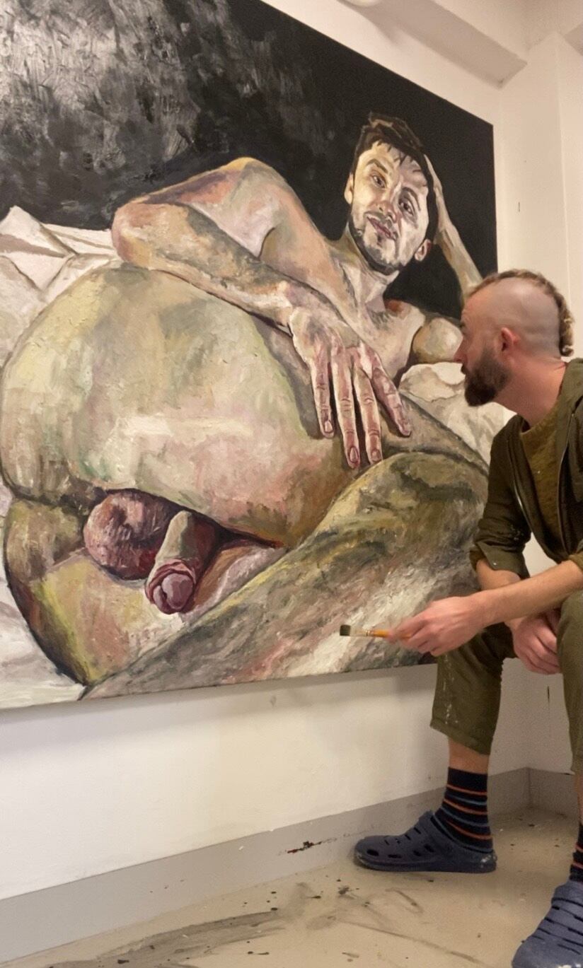 Ian Brennan sentado junto a una pintura expresiva de un hombre con mirada intensa y gesto vulnerable.