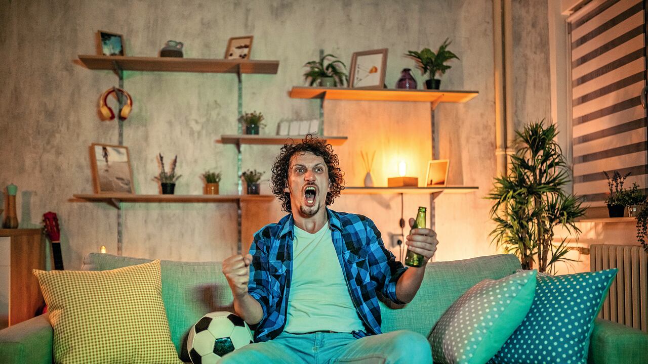 Handsome young man watching soccer match on TV at home in the evening and drinking beer. He is celebrating win of his team.