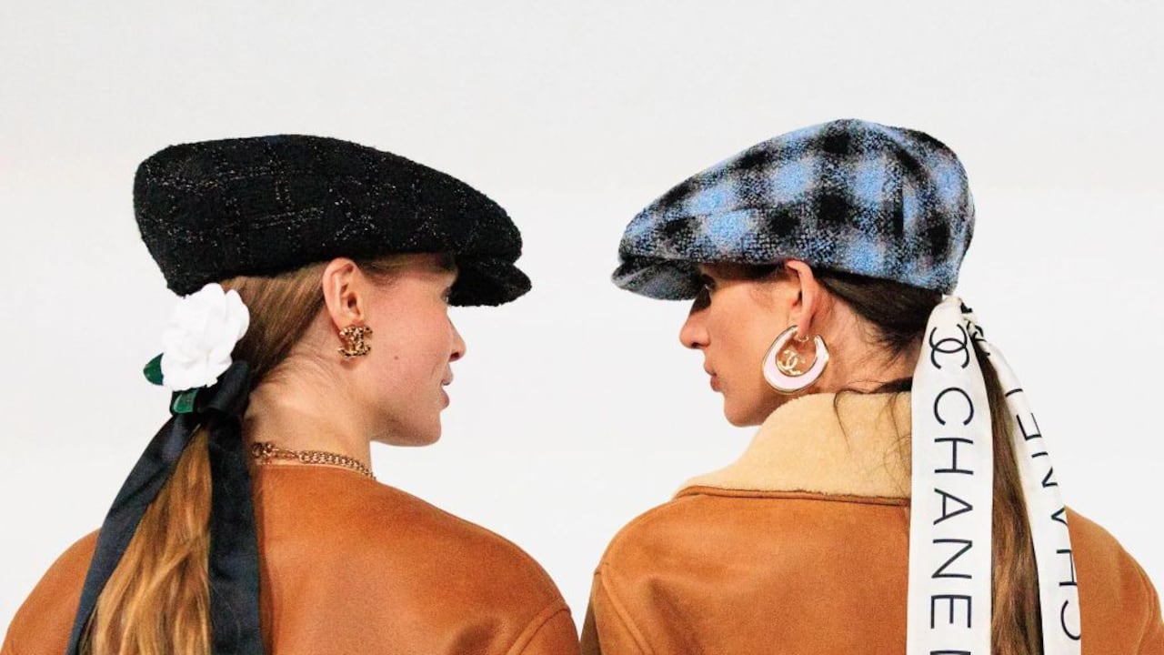 Dos mujeres de espaldas lucen boinas y abrigos en tonos tierra; una lleva una cinta con el logo de Chanel, evocando estilo clásico y sofisticación contemporánea.