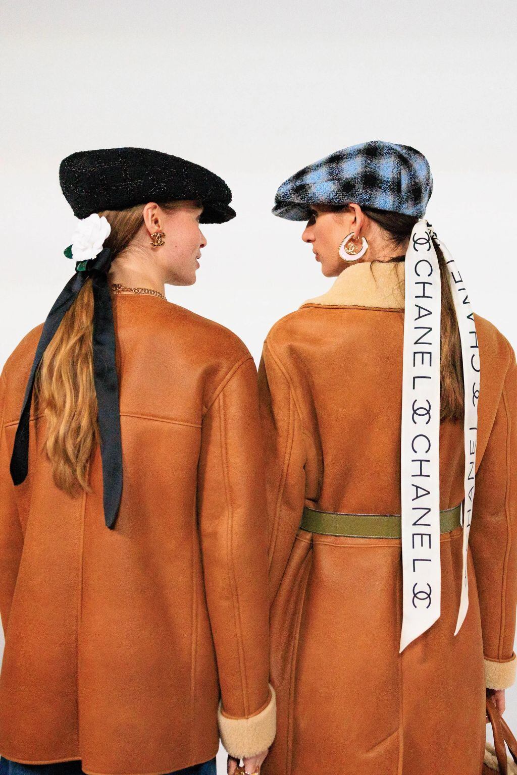 Dos mujeres de espaldas lucen boinas y abrigos en tonos tierra; una lleva una cinta con el logo de Chanel, evocando estilo clásico y sofisticación contemporánea.