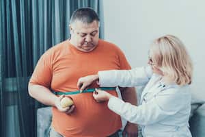 Hombre gordo con doctor en bata blanca en habitación gris. Mujer con centímetro de sastre. Concepto de dieta y salud. Hombre con Bulimia. Concepto de estilo de vida poco saludable. Hombre con sobrepeso. Paciente con Estómago.