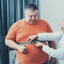 Hombre gordo con doctor en bata blanca en habitación gris. Mujer con centímetro de sastre. Concepto de dieta y salud. Hombre con Bulimia. Concepto de estilo de vida poco saludable. Hombre con sobrepeso. Paciente con Estómago.